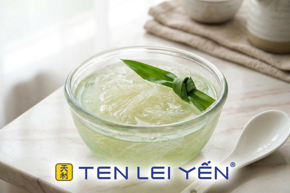 A finished double-boiled pandan bird's nest dessert in a glass bowl with pale jade-green broth, translucent bird's nest strands, and a knotted pandan leaf garnish beside a white porcelain spoon