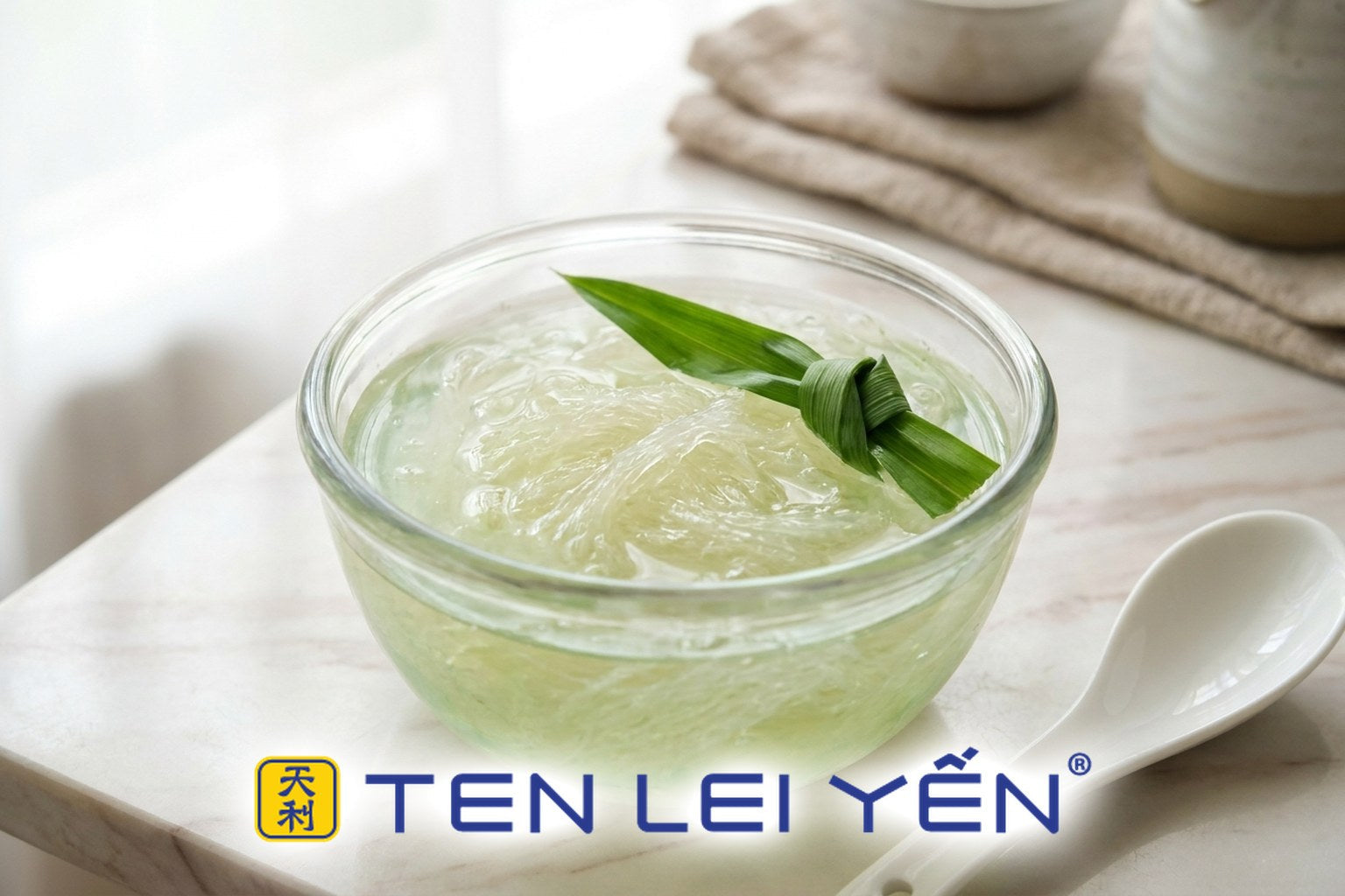 A finished double-boiled pandan bird's nest dessert in a glass bowl with pale jade-green broth, translucent bird's nest strands, and a knotted pandan leaf garnish beside a white porcelain spoon