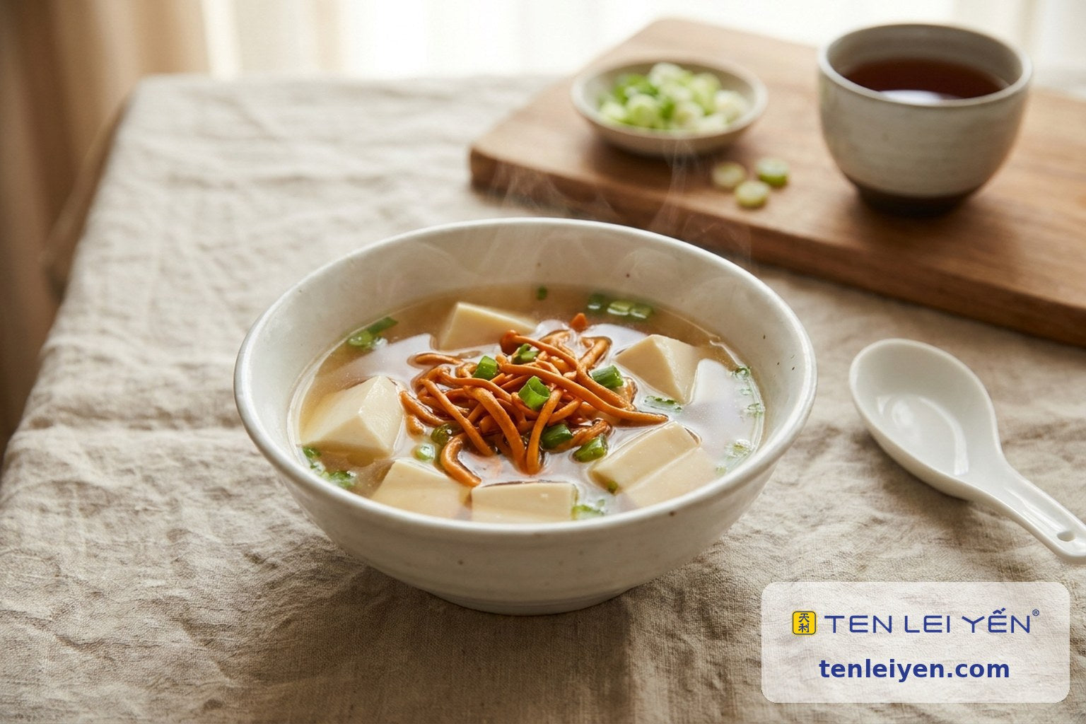 White porcelain bowl of Cordyceps militaris and silken tofu soup with clear broth and scallion beside a white porcelain spoon